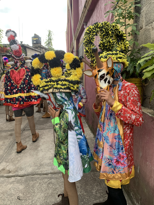 Eine Gruppe von Menschen in bunten Kostümen mit verschiedenen Gegenständen in der Hand, die vor einem Gebäude mit Bäumen und einem klaren blauen Himmel im Hintergrund stehen, wahrscheinlich an einem Karnevalsumzug teilnehmen.