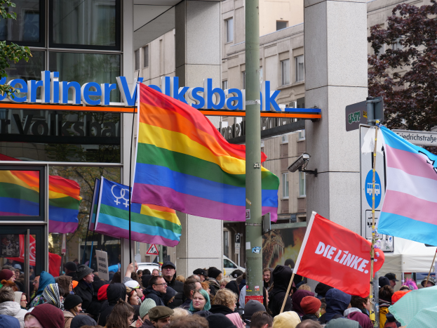 Eine große Gruppe von Menschen mit Fahnen und Schildern steht vor einem Gebäude, mit einem Mast im Vordergrund und Bäumen auf beiden Seiten, bei einer Gay-Pride-Veranstaltung in Berlin.