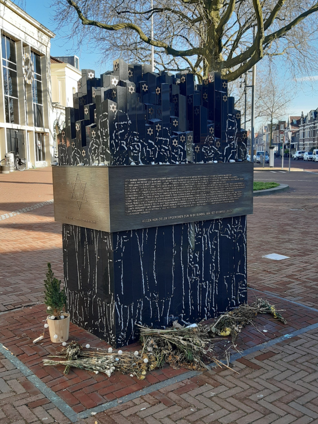 Denkmal in einem Stadtplatz, das Holocaust-Opfern gewidmet ist, mit einem Baum davor, einer Topfpflanze auf dem Boden, verstreuten trockenen Blättern und Gebäuden, Fahrzeugen, Pfählen und Bäumen im Hintergrund unter dem Himmel.
