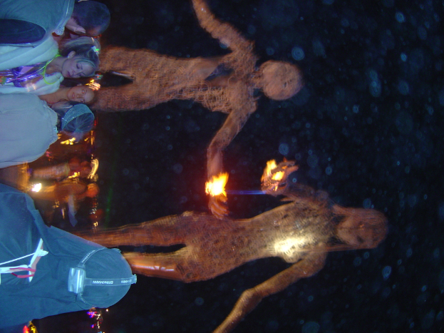 Menschen auf der linken Seite des Bildes mit brennenden Statuen dahinter und Lichtern im Hintergrund.