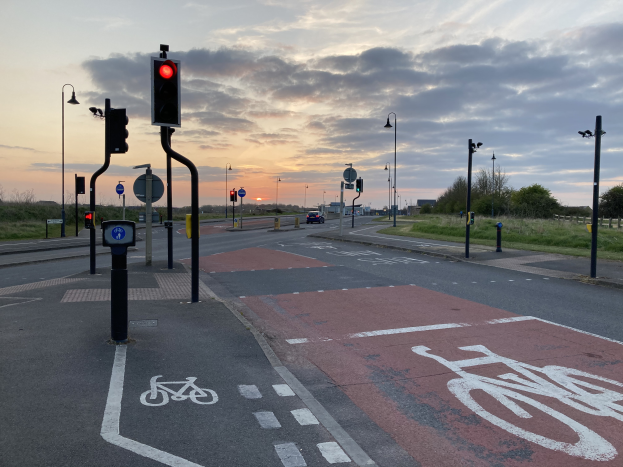 Ein Fahrradweg mit einer roten Ampel, Fahrzeuge auf der Straße, Laternenpfähle, Schilder, Gras, Bäume und ein bewölkter Himmel im Hintergrund.