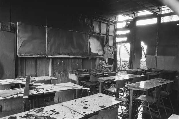 Schwarzes und weißes Foto eines Klassenzimmers mit verbrannten Wänden, zerbrochenen Fenstern und leeren Tischen und Stühlen.