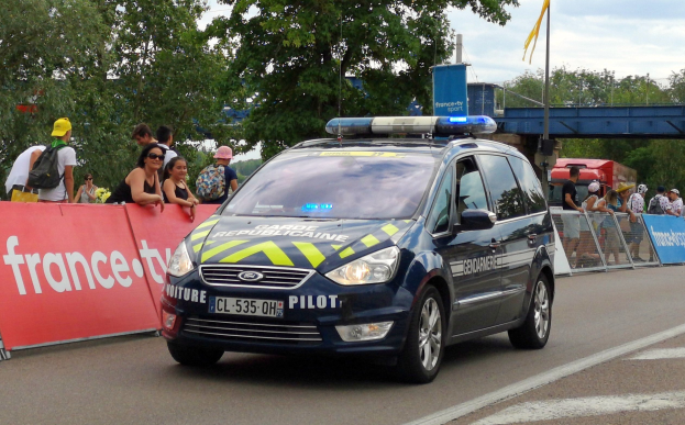 Ein Polizeiwagen fährt eine Straße entlang neben einer Menschenmenge, einige tragen Mützen und tragen Taschen, mit einem Banner links, Geländern mit Bannern dahinter und einem Hintergrund mit Bäumen, einer Brücke, einer Fahne an einem Mast und einem bewölkten Himmel.