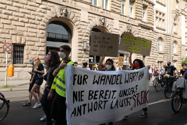 Eine Gruppe von Menschen marschiert auf einer Straße in einem Protest gegen die Anti-Kriegsbewegung, einige halten Schilder und andere fahren Fahrräder, mit einem historischen Gebäude, Bäumen und einer Straßenlaterne im Hintergrund.