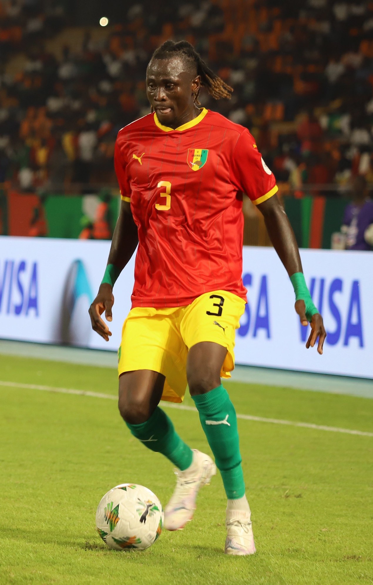 Ein Fuballspieler in einem rot-gelben Trikot, der einen Ball auf einem Feld mit Zuschauern und Werbetafeln im Hintergrund kickt.