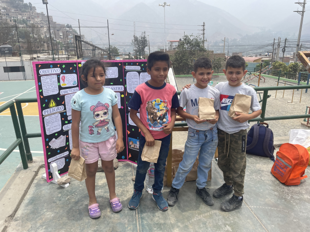 Gruppe von Kindern auf einem Basketballfeld mit Papiertüten in den Händen, mit Gegenständen auf dem Boden rechts und einer Tafel mit Text links, vor einem Hintergrund aus Zäunen, Pfosten, Straßenlaternen, Bäumen, Gebäuden, Hügeln und Himmel.