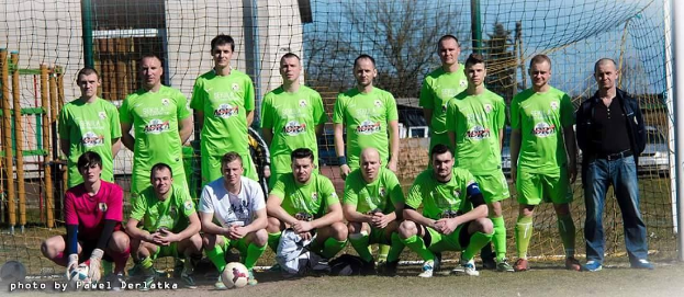 Gruppe von Männern in grünen T-Shirts auf einem Fußballfeld mit einem Ball und einem Tor, Bäume und Gebäude im Hintergrund.