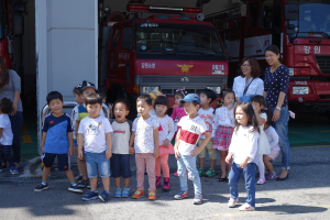 Eine Gruppe von Kindern vor mehreren Feuerwehren auf einer Feuerwache, einige tragen Mützen.