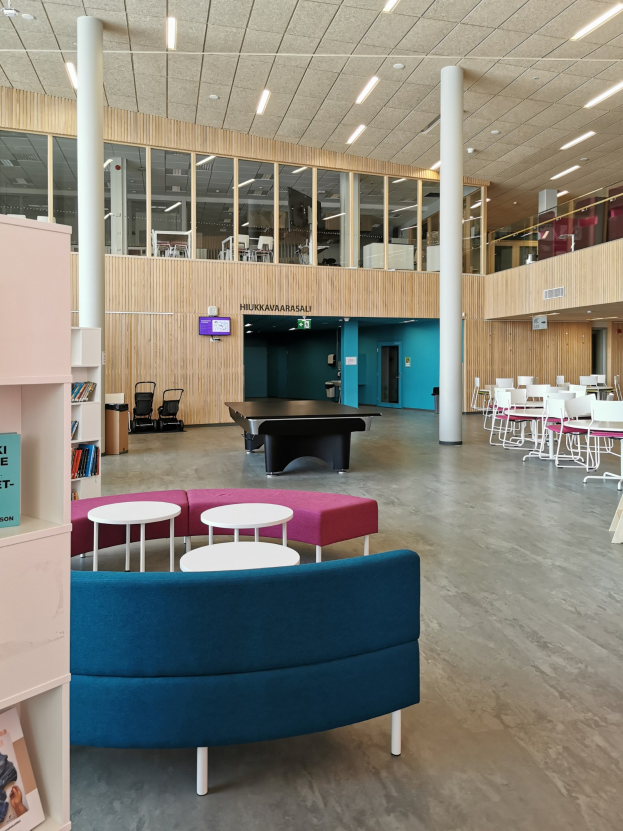 Großer Raum mit mehreren Tischen, Stühlen, Sofas, Hockern, Bücherregalen, Säulen, Deckenleuchten und einem Wandfernseher, der an ein Schulbibliotheksinnere erinnert.
