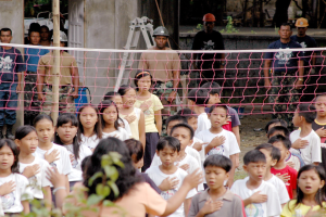 Eine Gruppe von Kindern vor einem Netz mit einer Frau im Vordergrund, während Menschen mit Helmen, eine Leiter, eine Wand und Bäume im Hintergrund zu sehen sind.