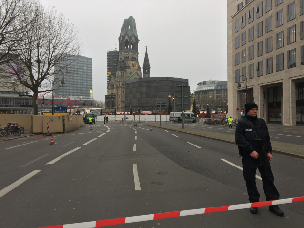 Ein Polizist in schwarzer Uniform und Mütze steht in einer Berliner Straße umgeben von Verkehrskegeln, Fahrrädern, Laternenpfählen, Bäumen und Gebäuden, mit einem Uhrenturm und einem klaren blauen Himmel im Hintergrund.