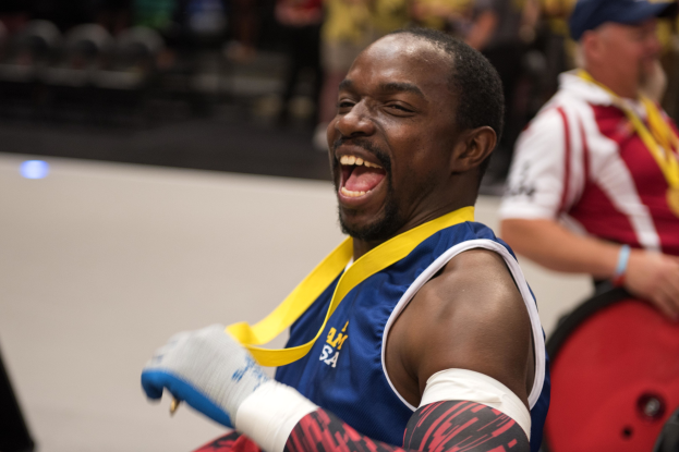 Ein Mann im Rollstuhl mit ausgestreckten Armen und einer Medaille um den Hals lächlt im Vordergrund, während im Hintergrund Menschen, Stühle und andere Gegenstände zu sehen sind.
