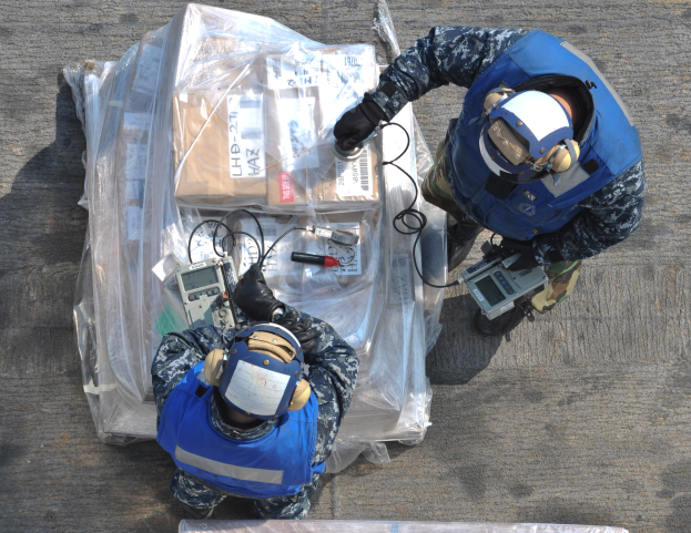 Zwei Männer in blauen Uniformen und Helmen arbeiten an einem großen Drohnenmodell und halten verschiedene Gegenstände in den Händen, vor ihnen liegt eine Plastikabdeckung auf dem Boden.
