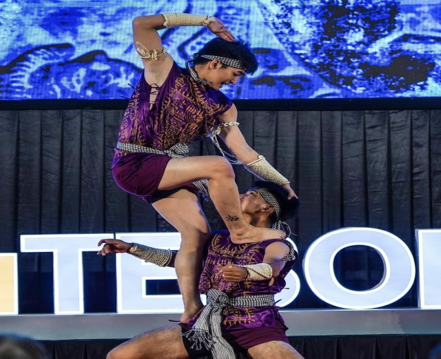 Zwei Performer in purpurfarbenen Kleidern führen einen akrobatischen Hebeflug auf der Bühne vor einem Publikum aus