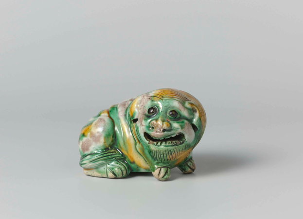 Eine kleine keramische Figur eines grünen und gelben lachenden Froschs mit einem verschmitzten Gesichtsausdruck auf einer weißen Oberfläche.