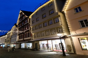 Headline in Rothenburg ob der Tauber, Deutschland, mit Weihnachtsbeleuchtung, Menschen und Fahrrad auf Kopfsteinpflasterstraße, flankiert von Gebäuden mit Fenstern unter einem sichtbaren Himmel.