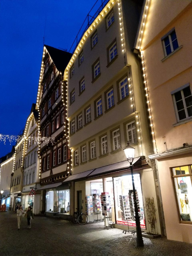 Headline in Rothenburg ob der Tauber, Deutschland, mit Weihnachtsbeleuchtung, Menschen und Fahrrad auf Kopfsteinpflasterstraße, flankiert von Gebäuden mit Fenstern unter einem sichtbaren Himmel.