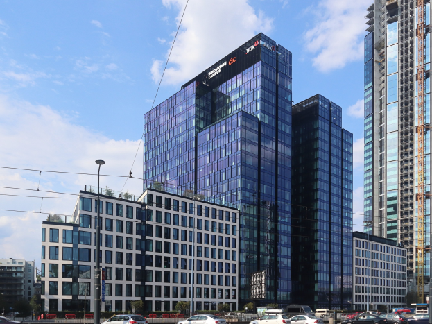 Zentrale der Deutschen Bank in Frankfurt, Deutschland, ein großes mehrstöckiges Gebäude mit zahlreichen Fenstern, umgeben von städtischer Infrastruktur wie Straßenlaternen, Elektrokabeln, Fahrzeugen, Grünflächen und einem bewölkten Himmel.