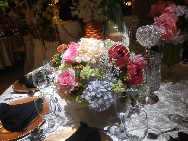 Ein formeller Esstisch mit Tellern, Besteck, Gläsern, Servietten und einem Blumenarrangement in der Mitte, umgeben von Stühlen und besetzten Tischen im Hintergrund.