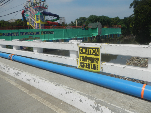Eine Brücke mit einem Warnschild, auf dem "Vorsicht temporäre Wasserleitung" steht, mit einem Geländer, Bäumen, Gebäuden, einer Wasserrutsche und einem bewölkten Himmel im Hintergrund.