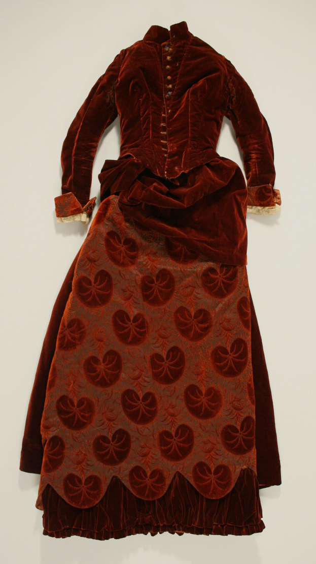 Ein rotes Kleid in einem Museum ausgestellt mit einem weißen Hintergrund, das ein hohes Dekolleté, lange Ärmel, einen weiten Rock, einen dezenten Glanz und detailreiche Spitzenverzierung zeigt.