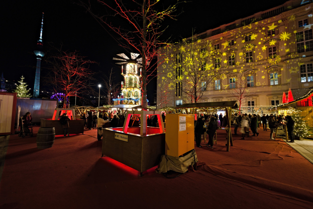 Ein geschäftiger Weihnachtsmarkt in Berlin, Deutschland mit Menschen um geschmückte Stände, Lichter, Bäume, Gebäude, Laternenmasten und einen Turm unter einem dunklen Himmel.