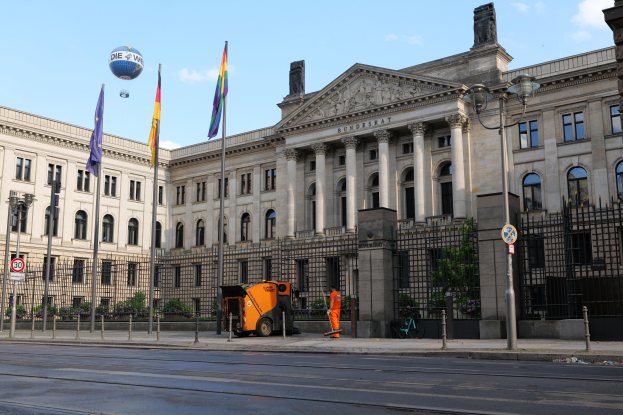 Das Reichstagsgebäude in Berlin, Deutschland, ein gro├čer Bau mit zahlreichen Fenstern, umgeben von Stra├čenmasten, Fahrzeugen, Passanten, Flaggen, Gittern, B├Ąumen und einem bew├Âlktem Himmel.