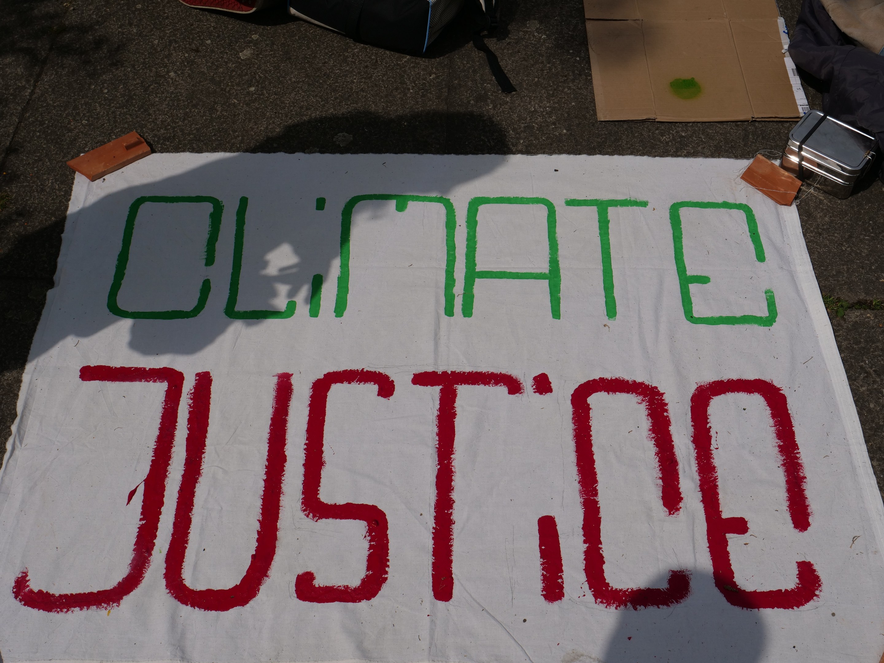 Weißer Banner mit "Klimagerechtigkeit"-Text auf dem Boden vor Pappe, einer Tüte und anderen Gegenständen.
