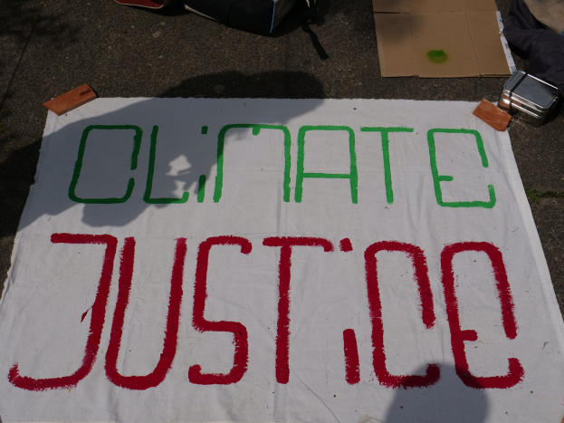 Weißer Banner mit "Klimagerechtigkeit"-Text auf dem Boden vor Pappe, einer Tüte und anderen Gegenständen.