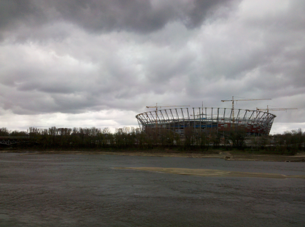 Das Olympiastadion in Kiew, Ukraine, umgeben von Bäumen und einer Brücke, mit bewölktem Himmel und sichtbarem Boden unten.