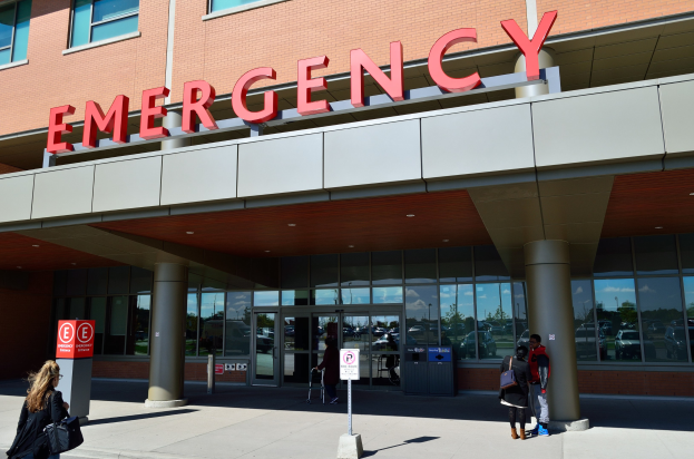 Eine Gruppe von Menschen geht vor einem Notfallgebäude mit Glaswänden, Säulen, einer Namensplakette, Schildern auf Pfählen und einem Mülleimer.