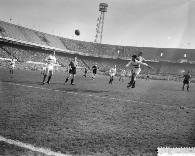 Eine Gruppe von Männern spielt Fußball auf einem Feld mit einem Stadion im Hintergrund, beleuchtet von Flutlicht unter einem klaren Himmel.