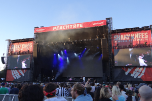 Eine große Menschenmenge steht vor einer hell erleuchteten Bühne auf dem Shaky Knees Music Festival, wo Musiker unter Bannern spielen, mit einem Geländer im Vordergrund und einem klaren blauen Himmel im Hintergrund.