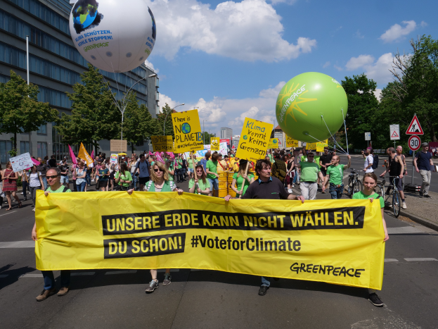 Eine Gruppe von Menschen marschiert mit einer gelben "Wähle für das Klima"-Plakette, Ballons, Schildern, Fahrrädern, Bäumen, Laternen und Gebäuden im Hintergrund unter einem klaren blauen Himmel.