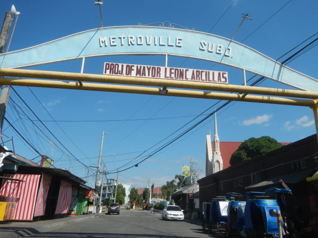 Bogen mit der Aufschrift "Metroville Subo - Projekt des Bürgermeisters Leon Arcillas" über einer Straße mit Fahrzeugen, Schuppen, Strommasten, Bäumen, Gebäuden und einem bewölkten Himmel.