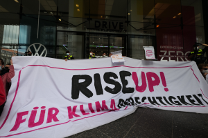 Eine Gruppe von Menschen mit Masken, die eine "Steh Auf"-Plakette und Schilder vor einem Glasgebäude halten, protestieren gegen eine Entscheidung der deutschen Regierung.