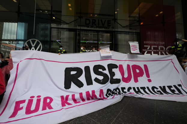 Eine Gruppe von Menschen mit Masken, die eine "Steh Auf"-Plakette und Schilder vor einem Glasgebäude halten, protestieren gegen eine Entscheidung der deutschen Regierung.