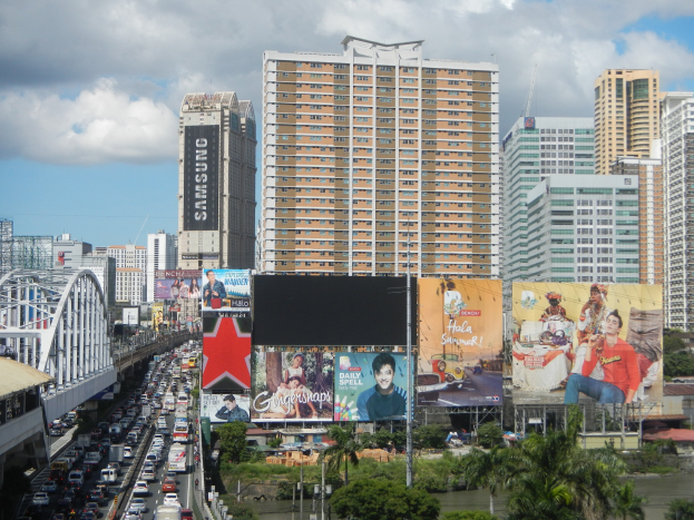 Eine belebte Straße mit starkem Verkehr, hohen Gebäuden, einer Brücke links, Bäumen, Masten, Plakaten und Wasser im Vordergrund, unter einem bewölkten Himmel.