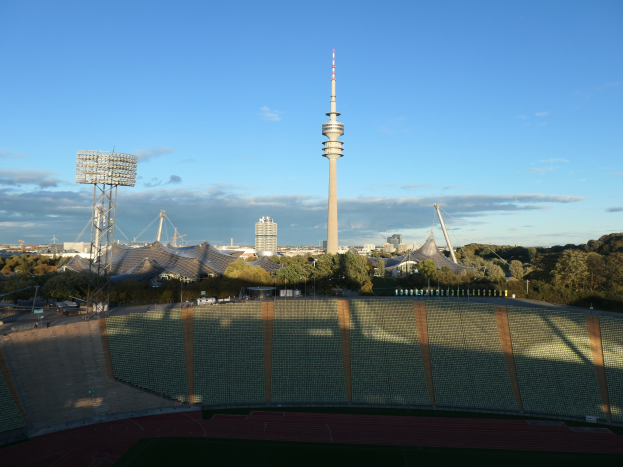 Das Olympische Stadion in Berlin, Deutschland, umgeben von Bäumen, Gebäuden und Lichtern, mit dem Fernsehturm im Hintergrund und einem bewölkten Himmel.