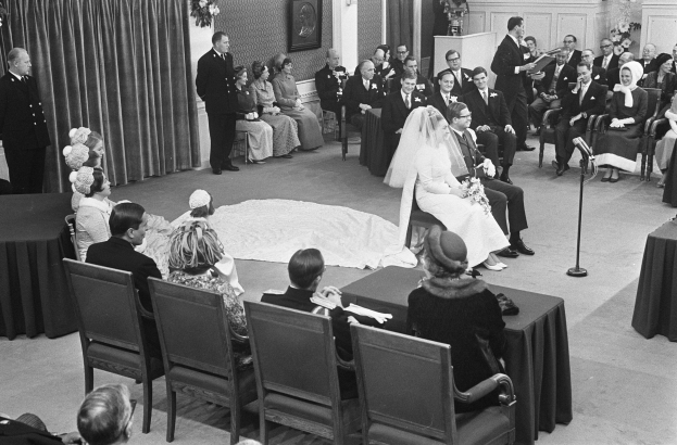 Schwarzes und weißes Foto einer Hochzeitszeremonie mit sitzenden Gästen, einem Mann, der in ein Mikrofon spricht, einer Frau mit einem Strauß, einem Podium mit Mikrofon, Blumenschmuck, einem Vorhang, einem gerahmten Foto an der Wand und einer Tür im Hintergrund.