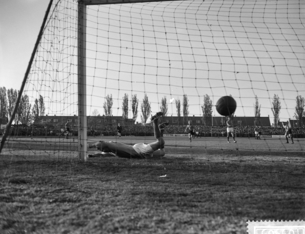 Ein Schwarz-Weiß-Foto eines Fußballspielers, der sich auf den Ball stürzt, vor einem Tor, mit anderen Spielern auf dem Boden, Bäumen, Gebäuden und einem klaren Himmel im Hintergrund.